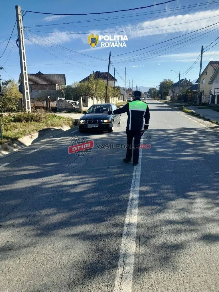 Un agent de poliție cu vestă reflectorizantă verde și neagră stă pe un drum asfaltat lângă o mașină neagră sub un cer senin.