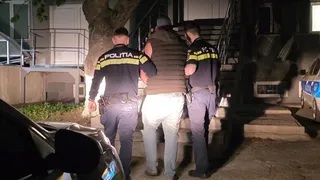 Trei polițiști în uniformă conduc un bărbat cu vestă maro și pantaloni deschiși pe o scară exterioară, noaptea.