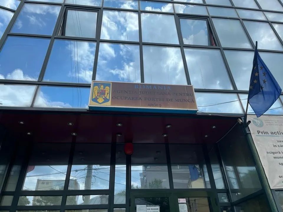 O clădire cu fațadă de sticlă reflectă cerul cu nori, având un panou informativ și un steag albastru cu stele galbene suspendat.