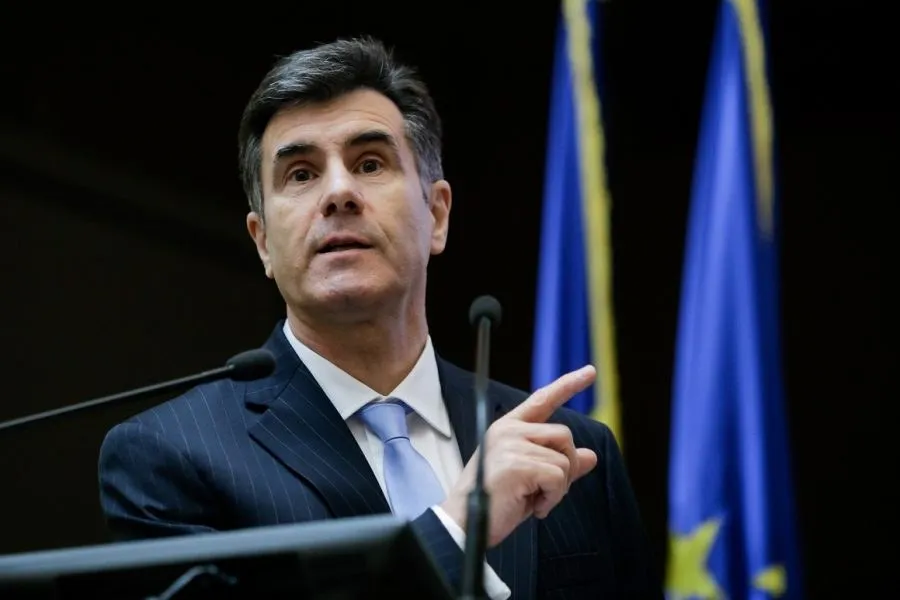 Un bărbat cu părul scurt și gri, purtând un costum în dungi și cravată albastră, ridică un deget în timp ce se află în fața unor microfoane.