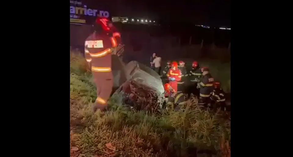 Mai mulți pompieri în uniforme reflectorizante stau lângă un vehicul overturnat într-un câmp de iarbă pe timp de noapte.