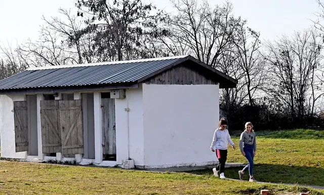 Două persoane merg pe un teren cu iarbă lângă o clădire mică, albă, cu acoperiș metalic înclinat și uși din lemn.