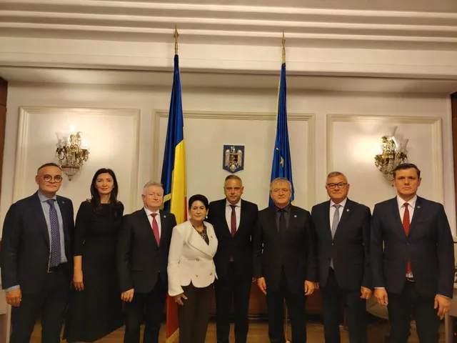 Opt persoane îmbrăcate în costume oficiale stau în rând în fața a două steaguri și a unui emblema pe un fundal alb cu panouri decorative.