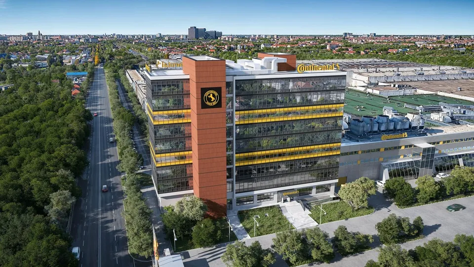 O vedere aeriană prezintă un clădire modernă cu fațadă de sticlă și panouri portocalii, situată lângă un drum și zone cu vegetație densă.