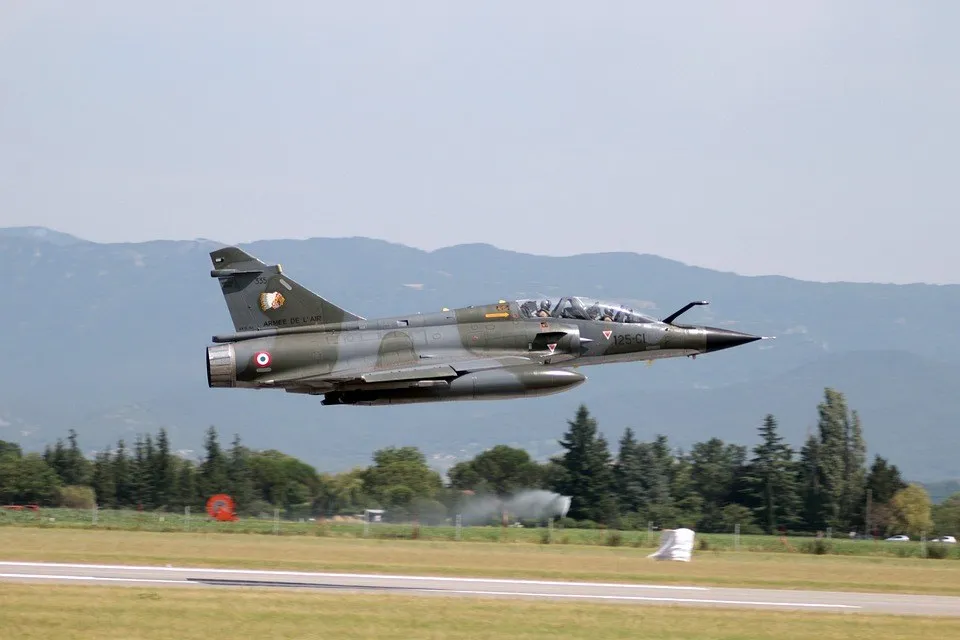 Un avion de vânătoare cu camuflaj verde și gri zboară deasupra unei piste de decolare lângă un lanț de munți.