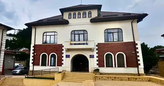 O clădire cu fațadă albă și secțiuni din cărămidă roșie prezintă ferestre cu rame negre și un balcon central deasupra unui hol de intrare.