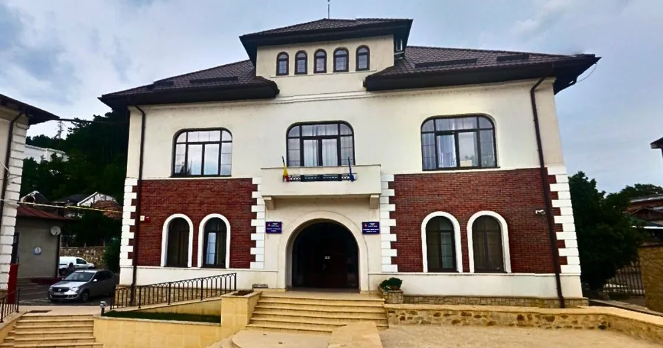 O clădire cu fațadă albă și secțiuni din cărămidă roșie prezintă ferestre cu rame negre și un balcon central deasupra unui hol de intrare.