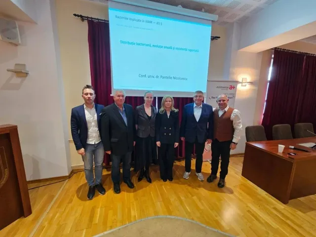 Șase persoane stau în rând în fața unui ecran de proiecție într-o încăpere cu podea de lemn și draperii roșii.