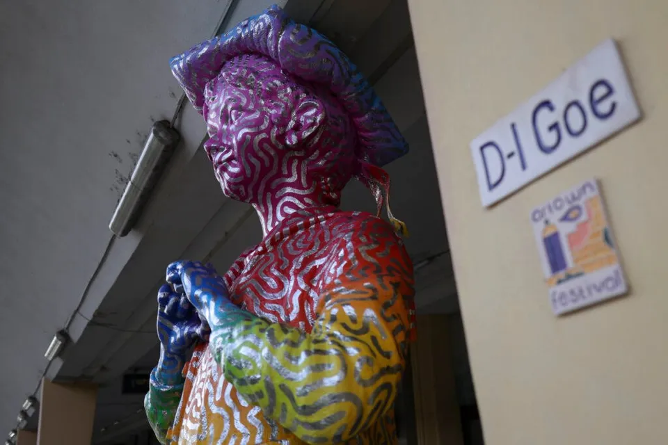 O statuie colorată în curcubeu, cu modele ondulate, stă lângă un perete pe care sunt afișate inscripția „D-l Goe” și un afiș ARTOWN.