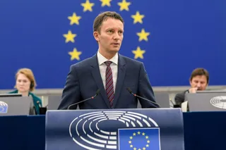 Un bărbat într-un costum închis la culoare și cravată roșie stă în spatele unui podium cu emblema Parlamentului European, în fața unui steag albastru cu stele galbene.