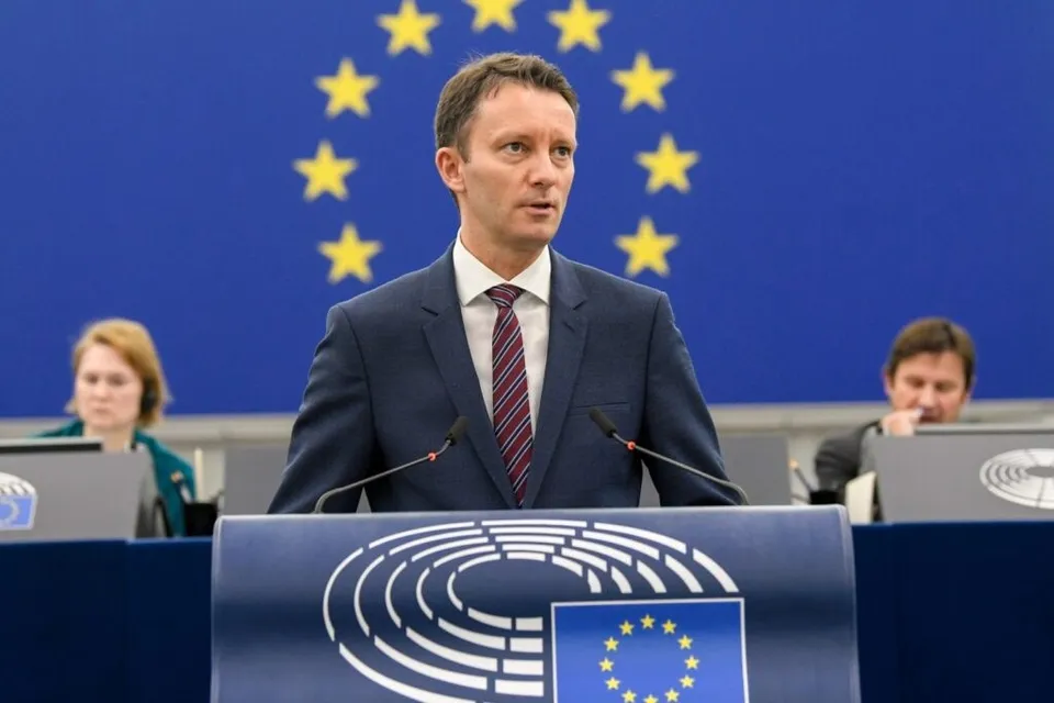 Un bărbat într-un costum închis la culoare și cravată roșie stă în spatele unui pupitru cu emblema Parlamentului European, în fața unui fundal albastru cu stele galbene.