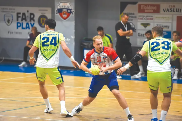 Trei jucători de handbal în echipamente colorate sunt pe un teren de lemn, unul dintre ei ținând o minge galbenă.