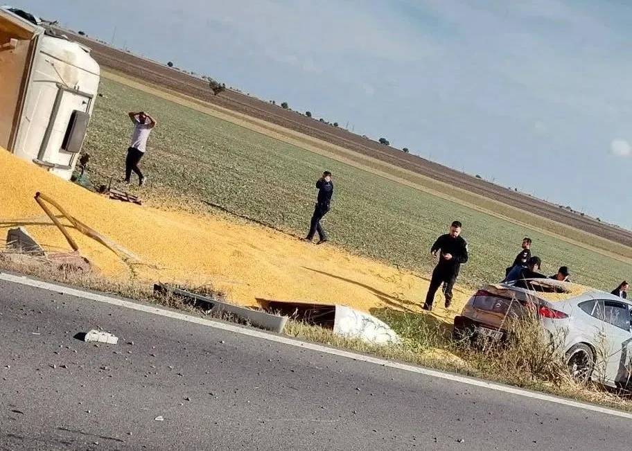 Un autoturism gri este parcat pe marginea unui drum lângă un câmp de cereale împrăștiate, în prezența câtorva persoane care stau în câmp.