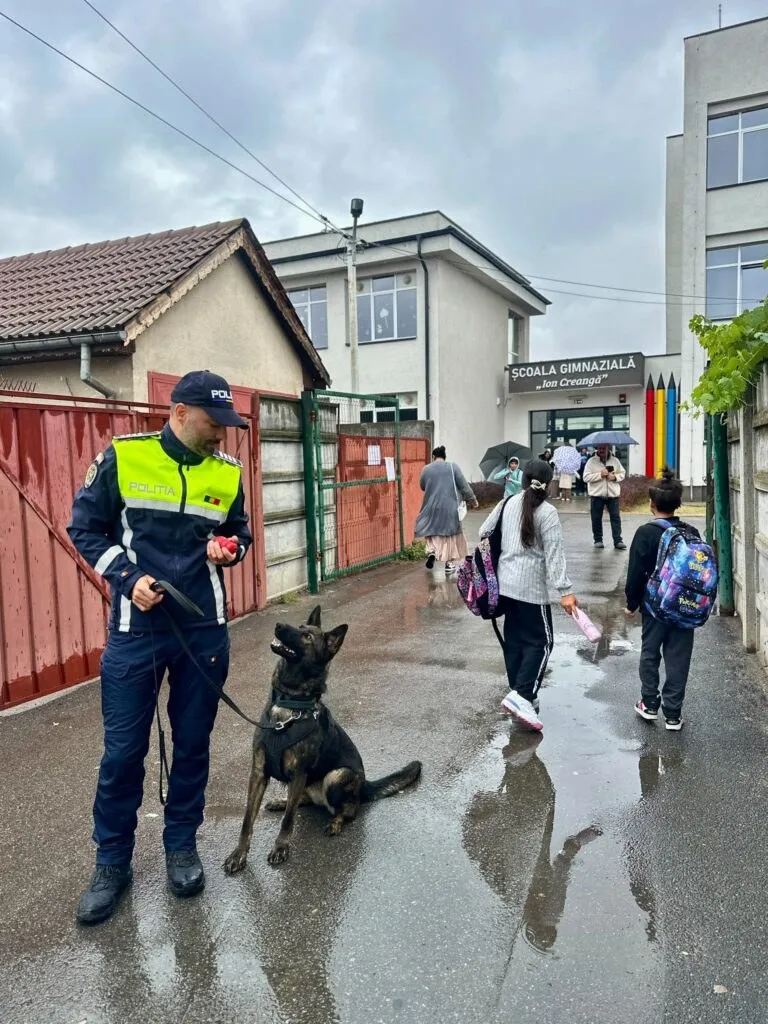 Un polițist cu un câine stă pe un trotuar umed lângă un grup de elevi care merg pe lângă o clădire școlară.
