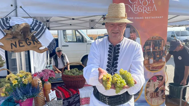 Un bărbat în costum popular ține struguri sub un cort, lângă un panou, coșuri și flori.