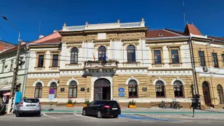 O clădire cu două etaje prezintă fațadă decorată, ferestre arcuite și un balcon central, având mașini parcate în fața sa pe un drum asfaltat.