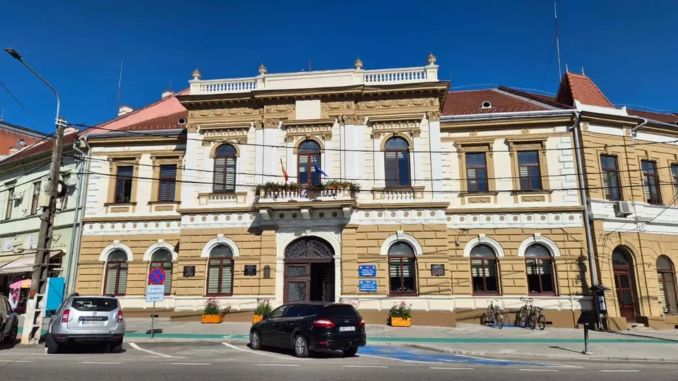 O clădire cu două etaje prezintă fațadă decorată, ferestre arcuite și un balcon central, având mașini parcate în fața sa pe un drum asfaltat.
