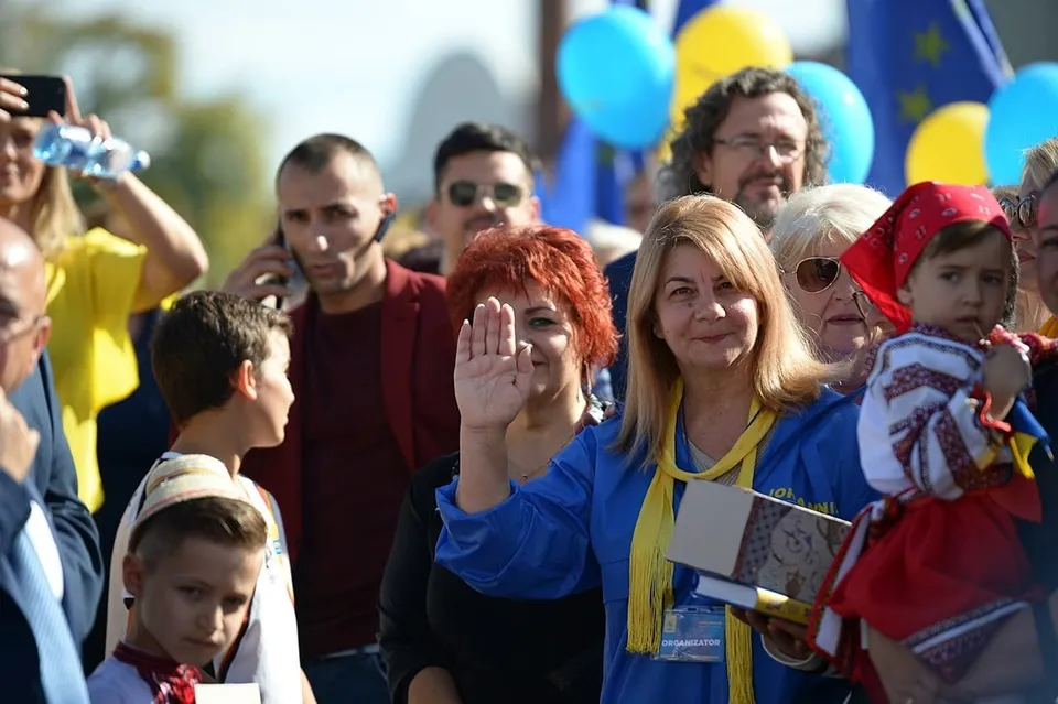 Un grup de persoane stă strâns la un eveniment în aer liber, printre care o femeie cu părul blond poartă un tricou albastru și un șal galben.