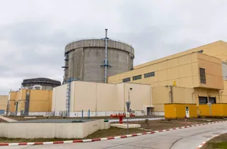Două clădiri industriale de culoare galbenă și gri sunt situate în fața unui cilindru masiv de beton sub un cer înnorat.