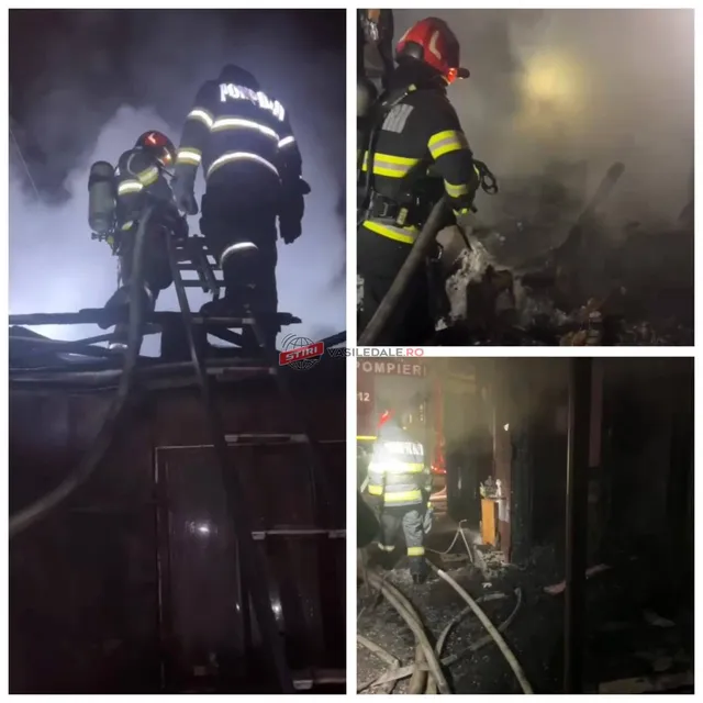 Pompieri cu echipament de protecție și echipamente de stingere lucrează noaptea lângă structuri de lemn în presence de fum.