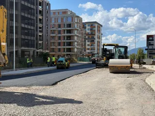 Mașini de construcții și lucrători cu veste reflectorizante se află pe un drum proaspăt asfaltat lângă clădiri înalte de apartamente sub un cer cu nori.