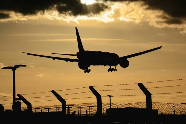 Silueta unui avion de pasageri în zbor pe fundalul unui cer la apus, deasupra unui gard de sârmă ghimpată.