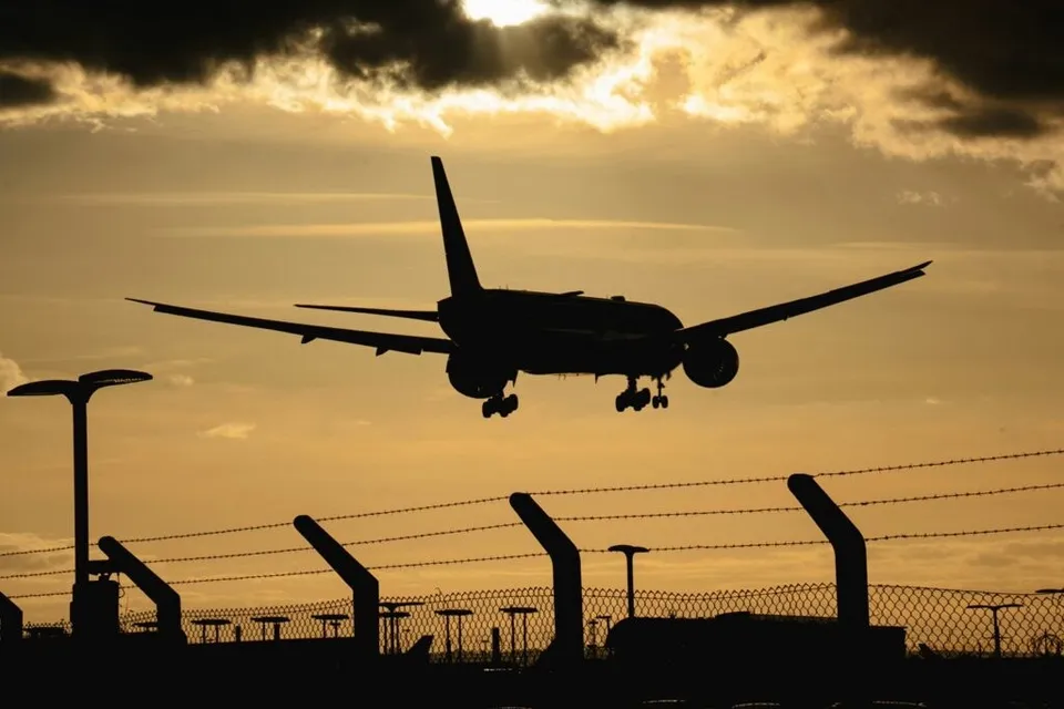 Silueta unui avion de pasageri în zbor pe fundalul unui cer la apus, deasupra unui gard de sârmă ghimpată.