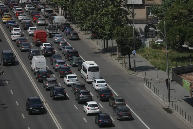 Mulți autovehicule de diverse culori și dimensiuni circulă pe mai multe benzi de circulație ale unui drum urban cu trotuare și copaci.