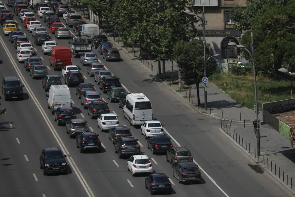 Mai multe vehicule de diverse dimensiuni și culori circulă pe o bandă de circulație asfaltată lângă un trotuar cu gard metalic și copaci verzi.