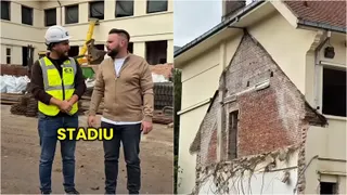 Un bărbat poartă vestă reflectorizantă galbenă și cască albă, stând lângă un alt bărbat într-o zonă de șantier lângă un perete de cărămidă expus.
