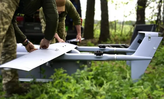 Două persoane în uniforme de culoarea verde manipulează un aparat aerian fără pilot de culoare gri aflat într-o zonă cu vegetație verde.