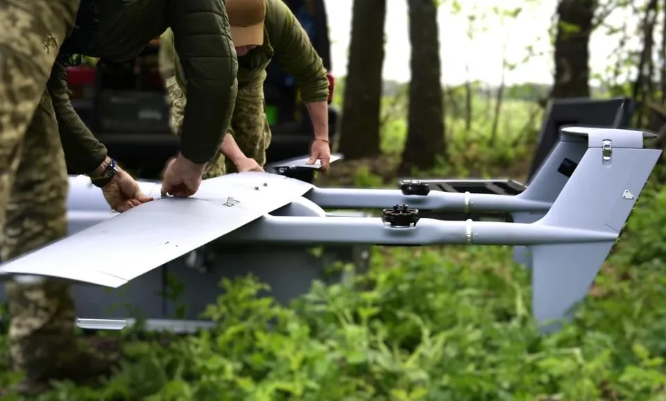 Două persoane în uniforme de culoarea verde manipulează un aparat aerian fără pilot de culoare gri aflat într-o zonă cu vegetație verde.