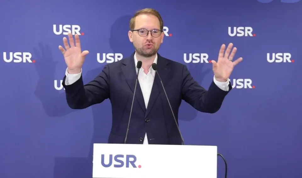 Un bărbat cu ochelari și sacou închis stă în spatele unui podium alb cu inscripția USR, având ambele mâini ridicate în lateral.
