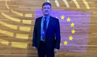Un bărbat într-un costum închis la culoare și cravată stă în picioare în fața unui perete din lemn cu un steag european proiectat pe el.