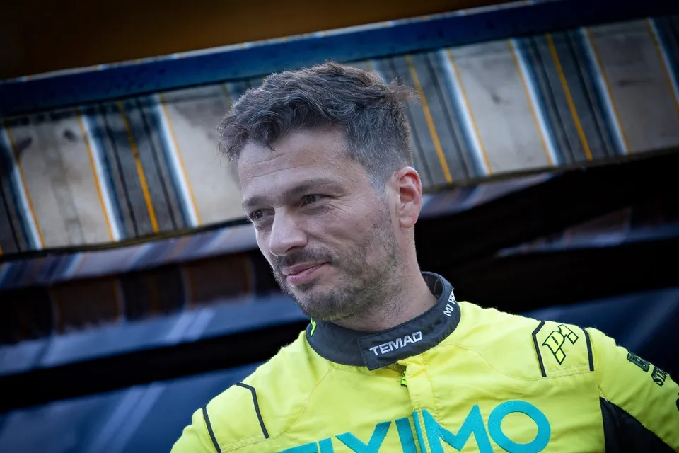 Un bărbat cu părul scurt și barbă stufoasă poartă un costum de racing galben cu detalii negre și albastre.