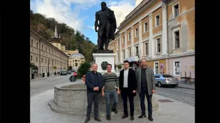 Patru bărbați stau în fața unei statui mari de culoare închisă, lângă o fântână rotundă, într-o zonă cu clădiri și un deal împădurit.