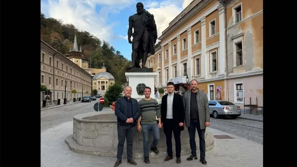 Patru bărbați stau în fața unei statui mari de culoare închisă, lângă o fântână rotundă, într-o zonă cu clădiri și un deal împădurit.