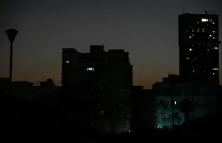 Siluetele unor clădiri înalte și ale unui stâlp de iluminat se ridică pe un cer închis, la apus.