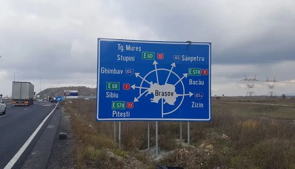 Un indicator rutier albastru cu săgeți și denumiri de orașe este amplasat pe marginea unei drumuri lângă un camion alb.