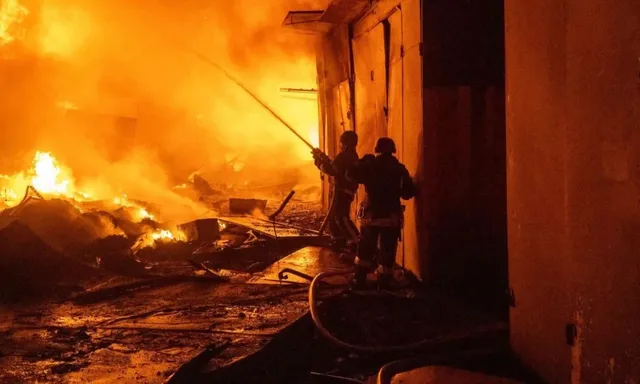 Două siluete de persoane stau lângă un furtun de apă în fața unui incendiu intens cu flăcări și fum dens.