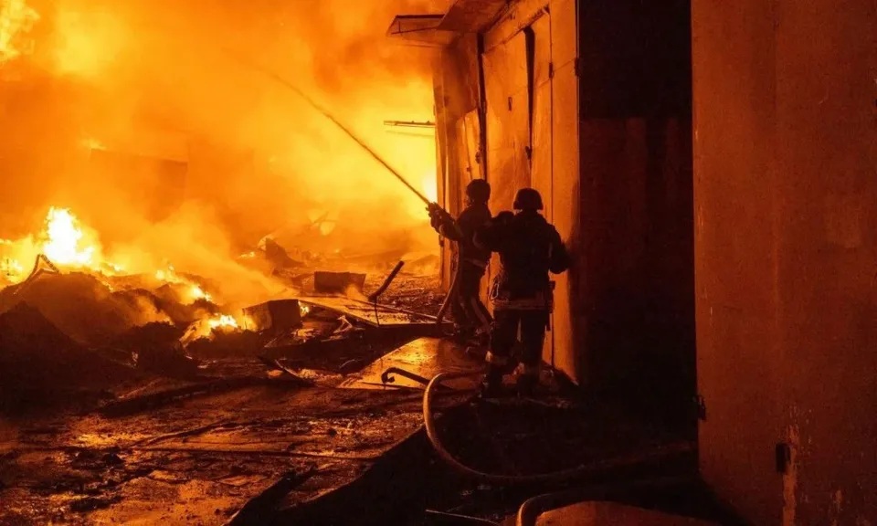 Două siluete de persoane stau lângă un furtun de apă în fața unui incendiu intens cu flăcări și fum dens.