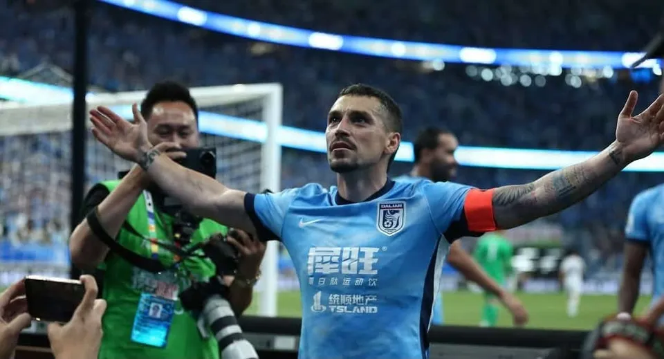 Un bărbat cu mâinile întinse poartă un tricou albastru pe un teren de fotbal, în timp ce un fotograf cu aparat foto stă în spatele lui.