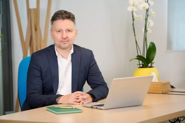 Un bărbat cu sacou închis și cămașă albă stă la o masă de lemn lângă un laptop și o plantă cu flori albe.