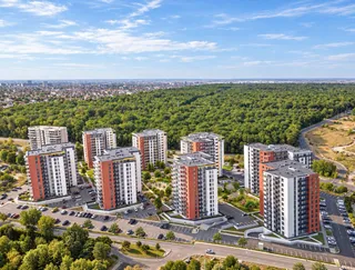 Mai multe clădiri de apartamente înalte, cu fațade albe și portocalii, sunt dispuse pe o suprafață verde lângă o pădure densă sub un cer senin.