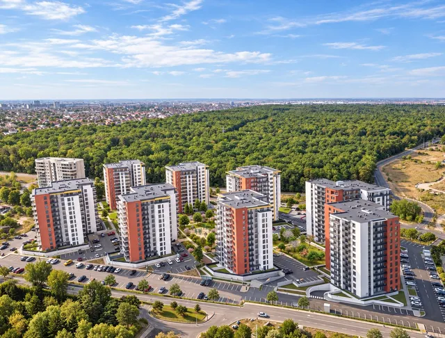 Mai multe clădiri de apartamente înalte, cu fațade albe și portocalii, sunt dispuse pe o suprafață verde lângă o pădure densă sub un cer senin.