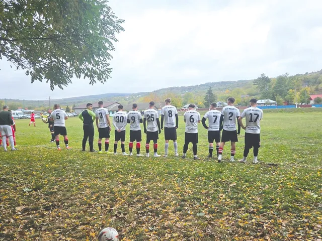O echipă de fotbaliști în echipamente albe și negre stă în linie pe un teren de fotbal acoperit de frunze căzute sub un cer înnorat.
