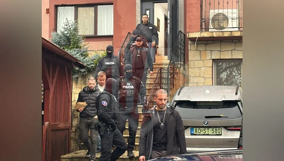 Mai mulți bărbați în haine negre și uniforme cu inscripția Politia stau în fața unei clădiri lângă o mașină de culoare gri.