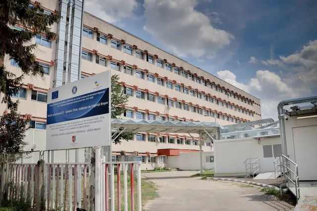 O clădire înaltă cu multe ferestre este vizibilă în spatele unui panou informativ alb și a unui gard metalic cu stâlpi albi și roșii.