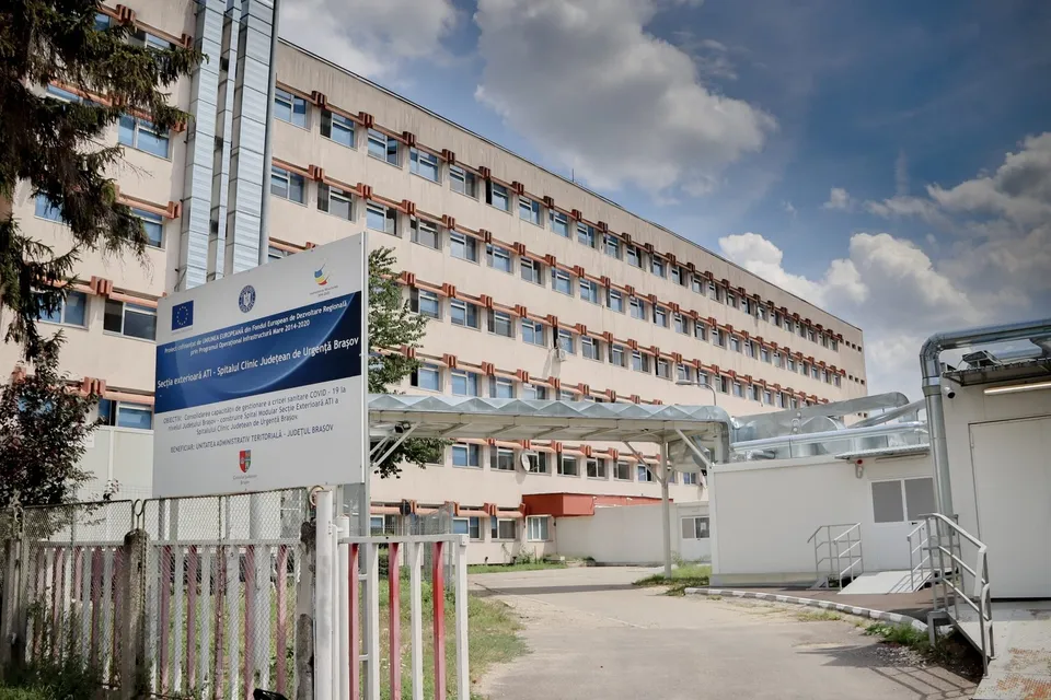 O clădire înaltă cu multe ferestre este vizibilă în spatele unui panou informativ alb și a unui gard metalic cu stâlpi albi și roșii.