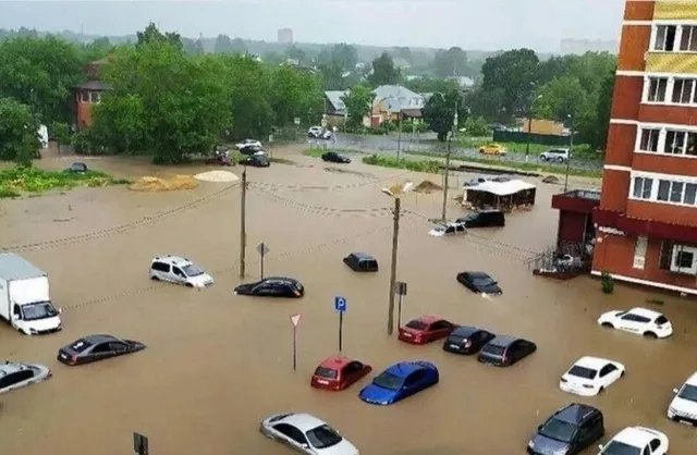 Mașini de diferite culori sunt parcate sau circulă pe un drum acoperit de apă de culoare maro, lângă clădiri și copaci sub un cer înnorat.